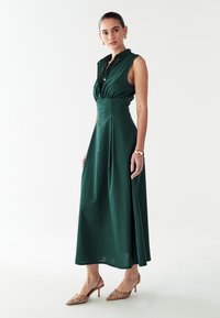 Grünes ärmelloses Kleid mit durchgehender Knopfleiste, tailliertem Schnitt und ausgestelltem Rock; aus leichtem Stoff gefertigt; kombiniert mit verzierten beigen Absätzen.