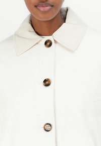 White coat with a corduroy collar, five large buttons featuring brown and metallic accents, and side pockets. Smooth fabric texture.