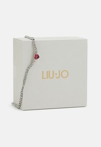 Bracelet en tone argenté avec des accents en cristal liés et une pierre précieuse rouge en forme de cœur, présenté sur une boîte grise avec du texte doré.