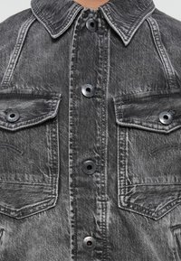 Primer plano de la parte frontal de una chaqueta de mezclilla negra descolorida que muestra el cuello, el cierre de botones y dos bolsillos en el pecho con solapa y costuras visibles.