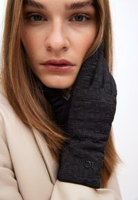 Černé prošívané rukavice s embosovaným textovým vzorem, s těsným střihem a staženým zápěstím, které má na sobě modelka s dlouhými vlasy.