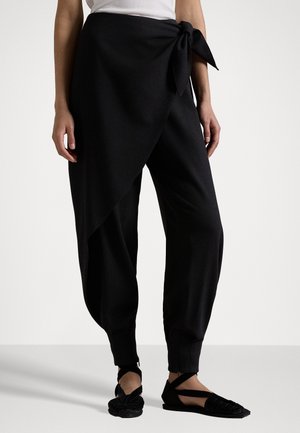 Zwarte, zachte stofbroek met een wikkeldesign en een zijstrikdetail. Heeft taps toelopende enkels en is gecombineerd met zwarte platte geweven schoenen.