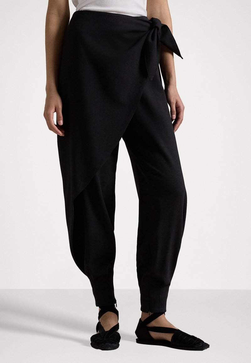 Zwarte, zachte stofbroek met een wikkeldesign en een zijstrikdetail. Heeft taps toelopende enkels en is gecombineerd met zwarte platte geweven schoenen.