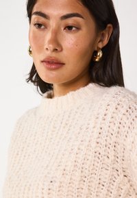 Maglione a maglia di un beige chiaro, con collo alto e una trama a tessitura larga. Il modello indossa grandi orecchini a cerchio dorati. L'attenzione è sui dettagli del maglione.