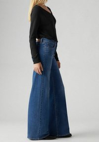 Niebieskie dżinsy o szerokich nogawkach i wysokim stanie, z pięcioma kieszeniami oraz gładką fakturą. Zestawione z czarną bluzką z długim rękawem.