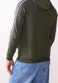 Sudadera verde oliva con tejido texturizado, franjas blancas a lo largo de las mangas y cintura ajustada, combinada con pantalones de mezclilla azul claro.