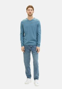 Helleblaue V-Ausschnitt-Jacke aus weichem Stoff, kombiniert mit hellblauen Jeans und weißen Sneakern. Die Jacke hat lange Ärmel und eine taillierte Passform.