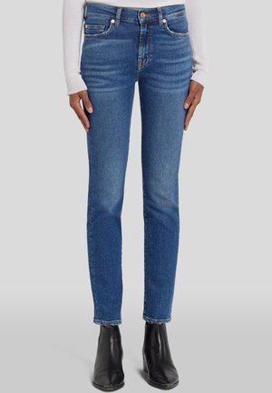 Dunne blauwe denimjeans hebben een middelhoge taille, vijf zakken, subtiele slijtplekken en worden gecombineerd met zwarte enkellaarzen.