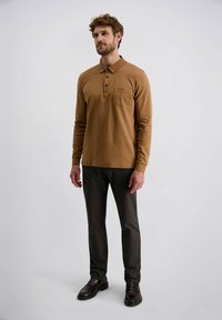Bruin polo shirt met lange mouwen en kraag, met een knoopsluiting van drie knopen en een borstzak, gecombineerd met donkergrijze pantalon en donkere schoenen.