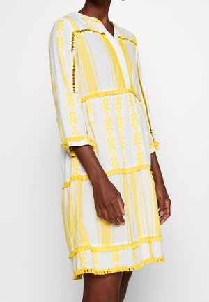 Kobieta ubrana w białą sukienkę do kolan z żółtymi pionowymi paskami i kwiatowymi wzorami, rękawy do ćwiartki oraz ozdobne frędzle.