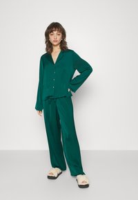 Ensemble de pyjama en satin teal composé d'une chemise à boutons avec col et d'un pantalon à taille élastique. Le mannequin porte des chaussons en fausse fourrure blancs.