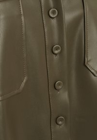 Camicia in ecopelle verde oliva con patta e quattro bottoni tondi, tasche appuntite e texture liscia con cuciture tono su tono.