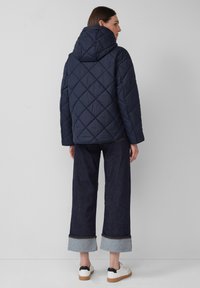 Marineblauwe gewatteerde hooded jas met een ontspannen pasvorm, voorzien van ruitpatronen, gecombineerd met wijdvallende donkere spijkerbroeken en witte sneakers.