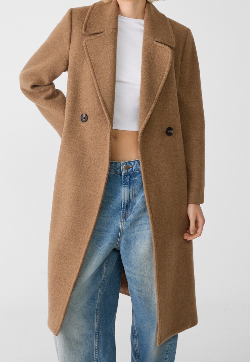 Femme portant un long manteau en laine camel sur un crop top blanc et un jean bleu ample, debout devant un fond uni.