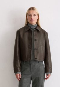 Braune Leder-Cropped-Jacke mit Kragen, Knopfverschluss und Fronttaschen. Getragen über einem grauen Rollkragenpullover mit glatter Textur.