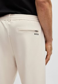 Beige broek met een gladde textuur, voorzien van een ritssluiting achterzak en het "BOSS"-logo in het zwart aan de rechterkant, dicht bij de tailleband.