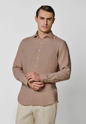 Mann steht mit verschränkten Händen, trägt ein hellbraunes Hemd mit Knöpfen und cremefarbene Hosen vor einem einfarbigen Hintergrund.