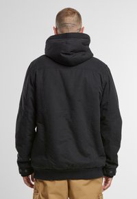 Chaqueta negra con capucha, de superficie texturizada, hombros caídos y puños con botones a presión. Presenta un ajuste relajado y un dobladillo elástico.