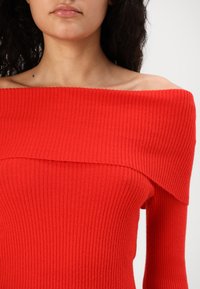 Maglione rosso a coste con design scoperto sulle spalle, maniche lunghe e finitura testurizzata. Il materiale sembra morbido e aderente.