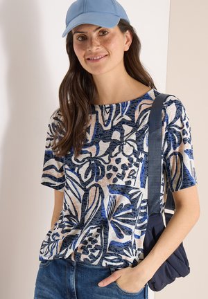 Frau mit langem braunem Haar, trägt ein blau gemustertes kurzärmeliges Hemd, eine hellblaue Kappe, Jeans und eine marineblaue Umhängetasche.