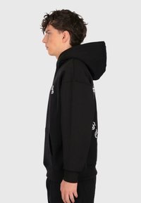 Jeune homme aux cheveux bouclés portant un sweat à capuche noir avec un texte blanc, montré de profil contre un fond lumineux et uni.