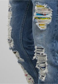 Jeans din denim albastru cu multiple rupturi orizontale, expunând un material colorat cu modele în interior.