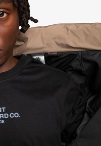 Sort t-shirt med logo, parret med en beige, polstret jakke, der er lynet op for at vise det sorte indvendige for. Teksturdetaljer på stoffet er synlige.