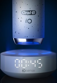 Brosse à dents électrique Oral-B iO avec un corps texturé bleu clair mat et une base en métal argenté, montée au-dessus d'une station de charge circulaire blanche affichant un minuteur.