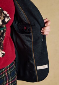Mano che tiene aperto un blazer scuro a spina di pesce che mostra la fodera nera con tasca interna ed etichetta, indossato sopra un maglione rosso con motivo cane e pantaloni a quadri.