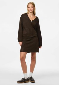 Brązowy sweter z dzianiny z zamkiem z przodu i kieszenią, w zestawie z pasującą brązową spódnicą. Modelka nosi czarne buty i białe skarpetki.