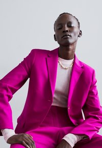 Blazer tailored rosa acceso abbinato a pantaloni e indossato su una maglietta rosa chiaro. L'insieme presenta dettagli minimi e una texture liscia.