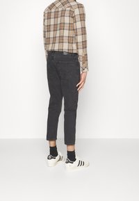 Jeans de mezclilla negra con corte slim, dobladillos ajustados y dos bolsillos traseros, combinados con una camisa de cuadros beige y zapatillas blancas con rayas negras.
