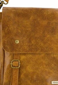 Sac messager en cuir marron texturé avec un rabat repliable, un bouton-pression en métal et une sangle réglable ; doté d'une boucle carrée et d'une étiquette de marque.