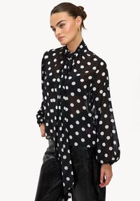 Blouse noire transparente à pois blancs, manches longues et col noué. Tissu texturé et poignets élastiques pour une coupe décontractée.