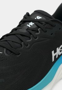 Imagem em close-up de um sapatilha desportiva preta com atacadores pretos, parte superior em malha respirável, logotipo branco "Hoka" e sola azul e branca.