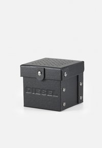 Boîte noire texturée avec couvercle, dotée d'un dessus perforé, logo "DIESEL" en relief et accents métalliques, y compris des boutons sur les côtés.