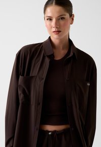 Braunes Hemd mit Knopfleiste, glatter Textur und einer Brusttasche. Getragen über einem enganliegenden, braunen gerippten Crop-Top.