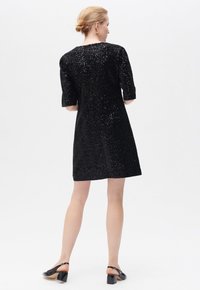 Vestido negro con mangas cortas, tejido texturizado, corte a la altura de la rodilla y un ligero brillo. Llevado con zapatos de talón descubierto y tono de piel suave.