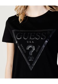 Must t-särk musta puuvillast, millel on suur läikiv must kolmnurga logo ja tekst "GUESS U.S.A. WASHED JEANS" koos küsimärgi. 
