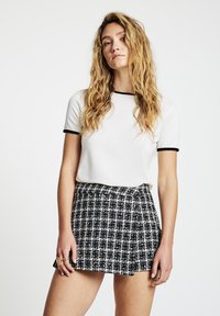 Weißes kurzärmeliges Oberteil mit schwarzem Besatz, kombiniert mit einem karierte Minirock in Schwarz, Weiß und Gold. Strukturierter Stoff.