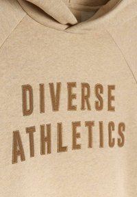 Zbliżenie beżowego hoodie z brązowym haftowanym napisem "DIVERSE ATHLETICS" na klatce piersiowej, ukazujące strukturę tkaniny i detale szycia.