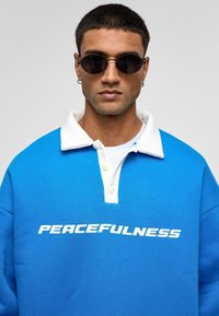 Blauwe sweatshirt met witte kraag en voorop de opdruk "PEACEFULNESS." Heeft een lichtgewicht materiaal en een losse pasvorm, gecombineerd met ronde zwarte zonnebril.