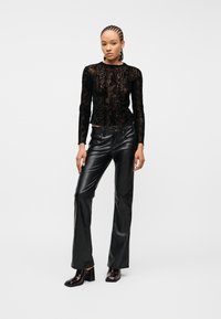 Γυναίκα με πλεξούδες που φοράει μαύρο διαφανές μακρυμάνικο τοπ με μοτίβο φιδιού, μαύρο δερμάτινο παντελόνι και μαύρα μποτάκια με τακούνι, στέκεται σε λευκό φόντο.