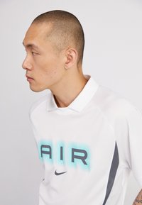 Biela polokošeľa s nápisom "AIR" v akvamarínovom gradiens, logom Nike a šedými akcentmi. Hladká textúra s golierom.