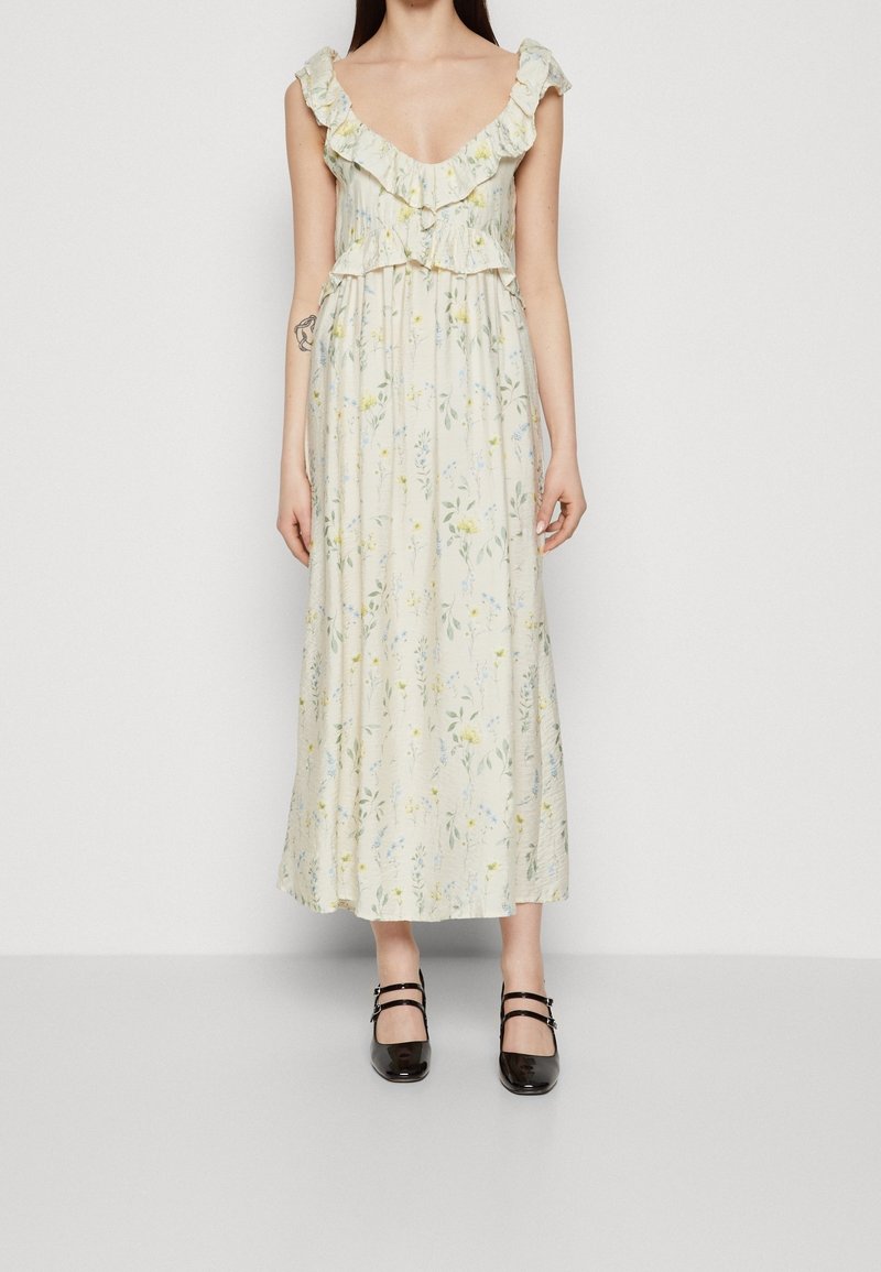 Robe maxi florale en crème, avec des manches volantées et un corsage à volants. Imprimé incluant des fleurs jaunes et bleues. Associée à des chaussures noires.