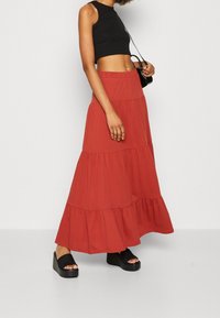 Jupe maxi rouge à volants avec une taille élastique, assortie à un crop top sans manches noir. Le modèle porte des sandales à plateforme noires.