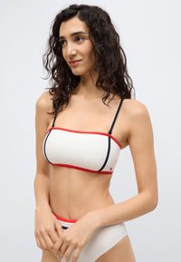 Σετ μπικίνι με κρέμ τοπ που διαθέτει κόκκινες και ναυτικές λεπτομέρειες, χωρίς τιράντες, και το κάτω μέρος σε κρέμ με ανάλογο διακοσμητικό περίγραμμα. Λείο υλικό.