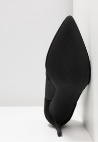 Schwarzer High Heel mit spitzem Zehenbereich und glatter Oberflächenstruktur. Die Sohle ist aus Gummi mit horizontalen Rillen für besseren Grip.