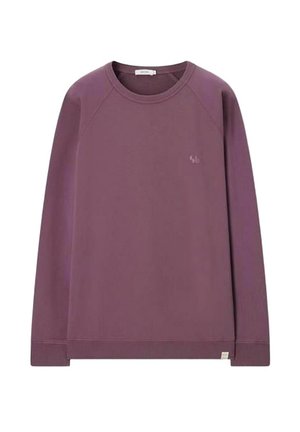 Homme debout, mains dans les poches, portant un sweat mauve, un short beige et des chaussures de running grises sur fond beige.