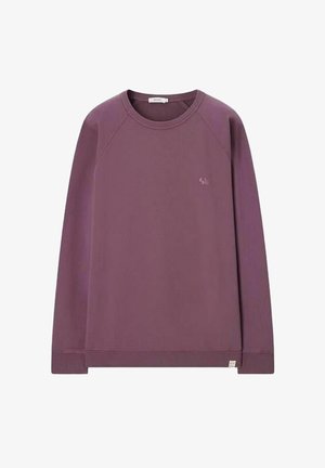Homme debout, mains dans les poches, portant un sweat mauve, un short beige et des chaussures de running grises sur fond beige.